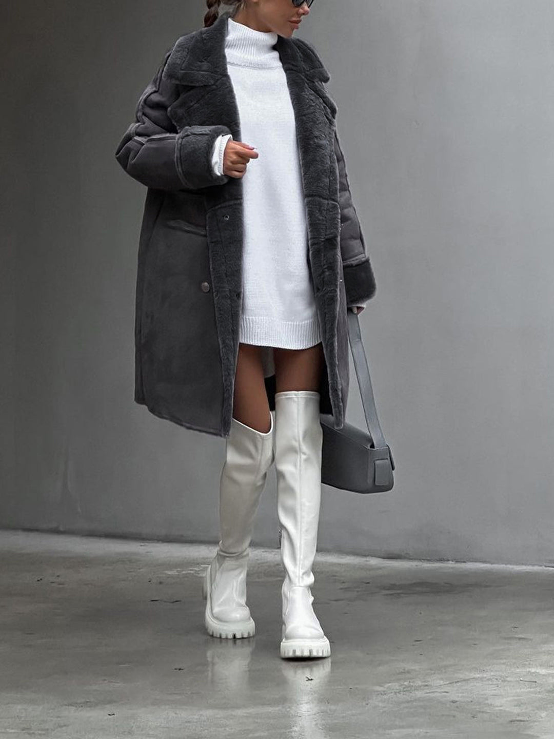 Person wearing a long dark coat, white turtleneck, white knee-high boots, and holding a black handbag against a gray background.
