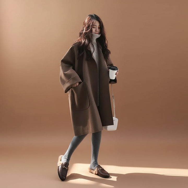 Woman in a brown coat holding a coffee cup against a white wall.